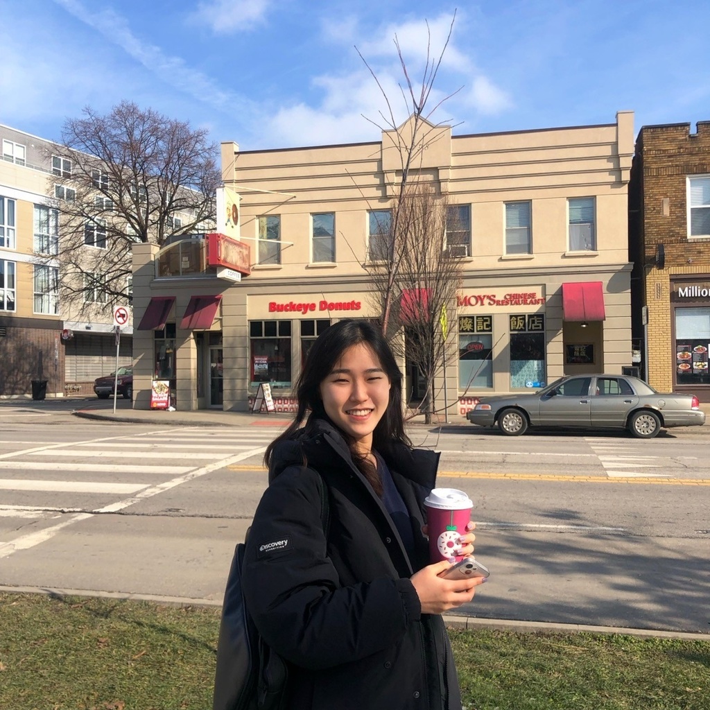 A photo of Danica walking on a street. Danica is holding a coffee cup and wearing black outfit. 