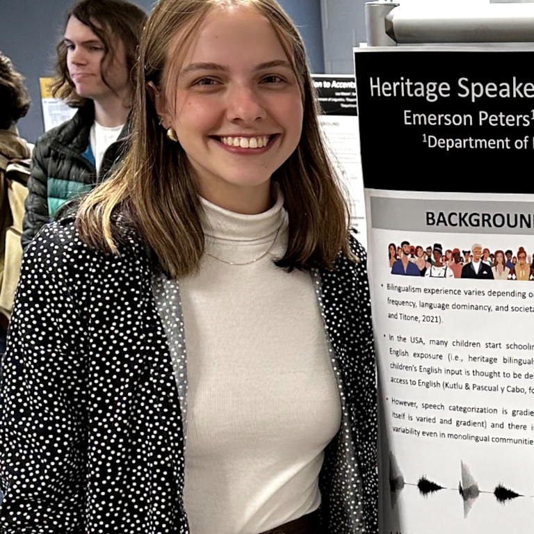 A picture of Emerson while presenting her scientific poster. Emerson is wearing a white shirt and gray cardigan. 