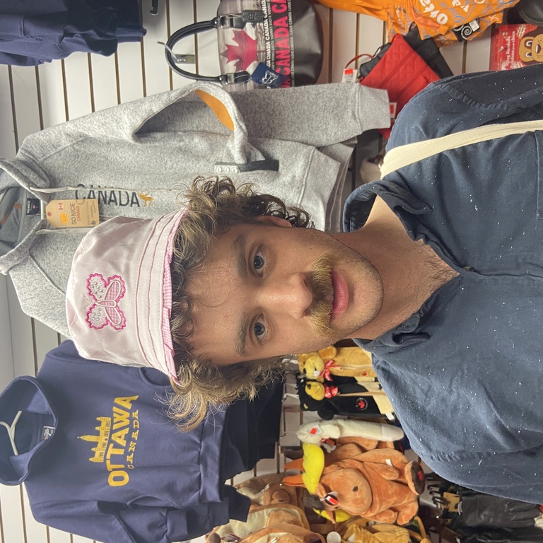 A picture of Jake in Ottawa, Canada in a souvenir shop wearing a pink hat while standing in front of a canada sweatshirt.