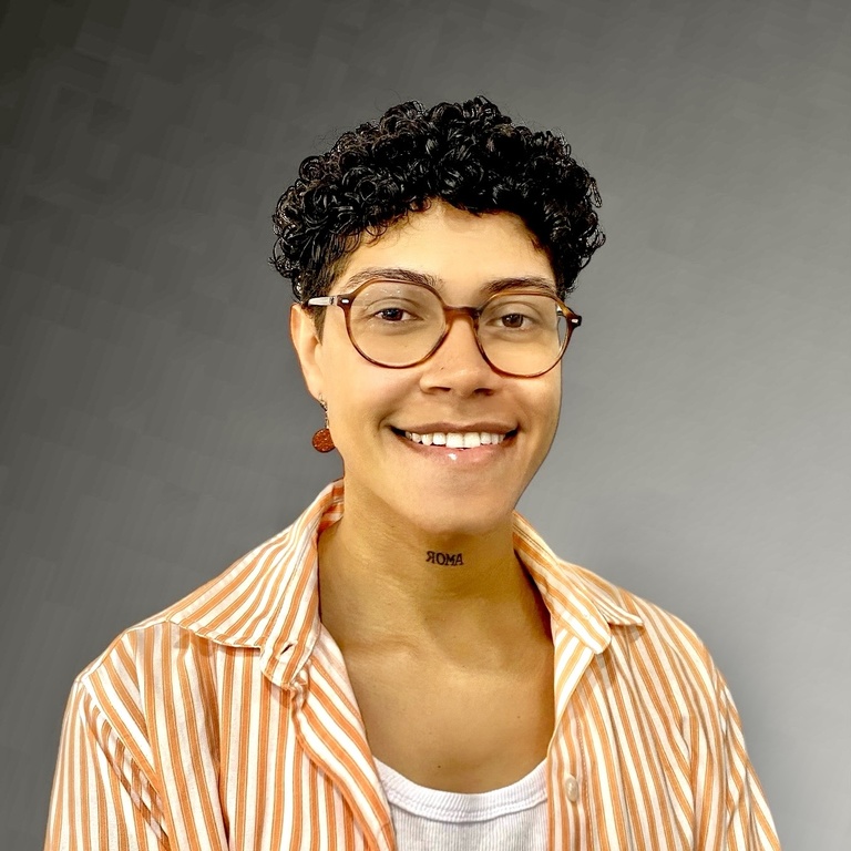 A picture of Bremdellin that was taken in front of a gray wall. Bremdellin is wearing an orange shirt. 