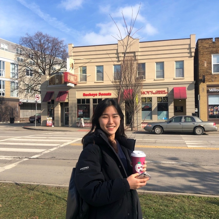 A photo of Danica walking on a street. Danica is holding a coffee cup and wearing black outfit. 